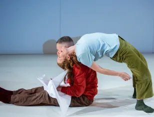 Eine Person sitzt mit brauner Hose und rotem Pullover auf dem Boden, eine andere lehnt mit blauem Oberteil und grüner Hose nach hinten über sie.