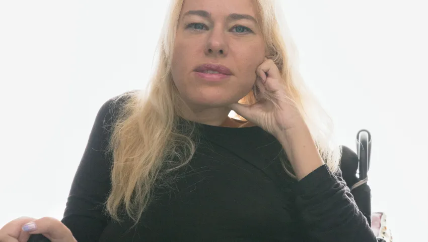 Portät einer Person mit langen blonden Haaren und dunkler Kleidung, die in einem Rollstuhl sitzt.