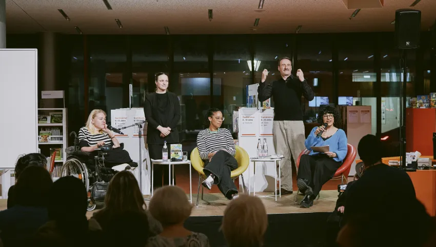 Ein Podium mit drei Frauen im Gespräch. Zwischen Ihnen steht ein Gebärdensprachendolmetscher und eine Gebärdensprachendolmetscherin