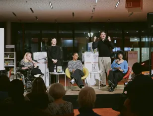 Ein Podium mit drei Frauen im Gespräch. Zwischen Ihnen steht ein Gebärdensprachendolmetscher und eine Gebärdensprachendolmetscherin