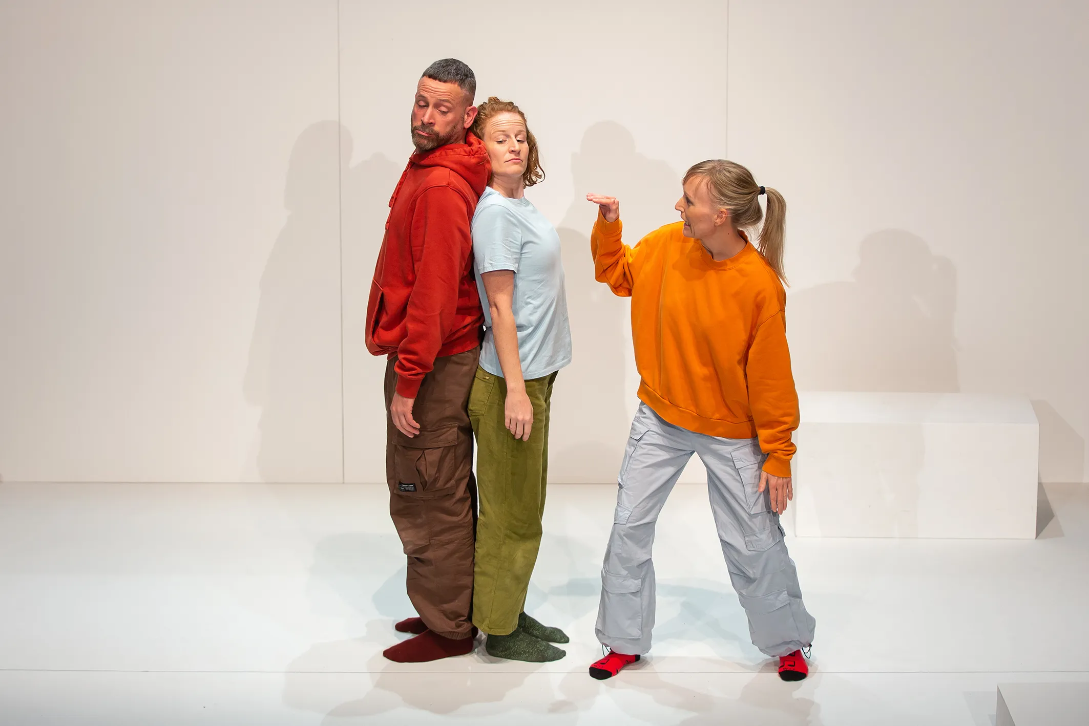 Zwei Personen stehen Rücken an Rücken zueinander, eine andere in orangenem Pullover gestikuliert mit ihrer Hand in ihre Richtung.
