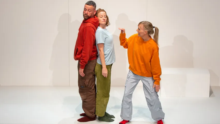 Zwei Personen stehen Rücken an Rücken zueinander, eine andere in orangenem Pullover gestikuliert mit ihrer Hand in ihre Richtung.