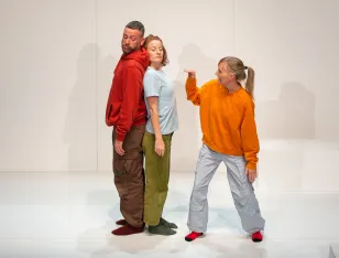 Zwei Personen stehen Rücken an Rücken zueinander, eine andere in orangenem Pullover gestikuliert mit ihrer Hand in ihre Richtung.