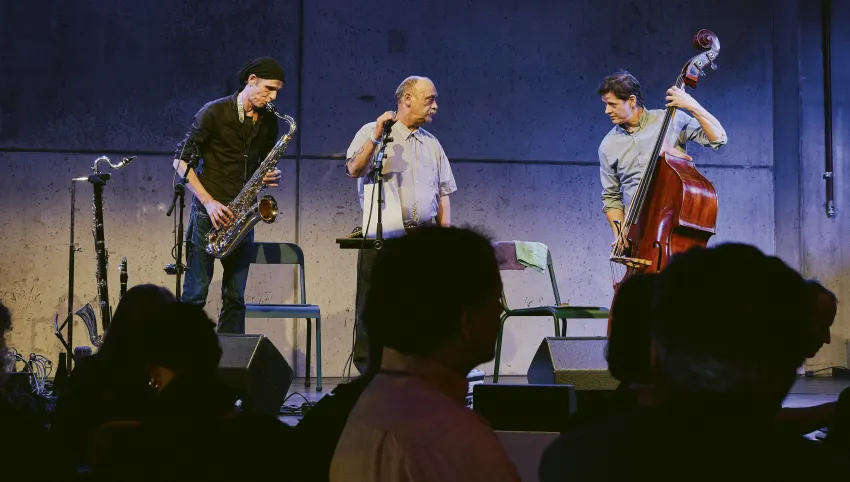 Momentaufnahme des Auftritts von O'Chavo Pretzlie bei der Brechtnacht. Ein Saxophonist, ein Sänger, ein Kontrabassist