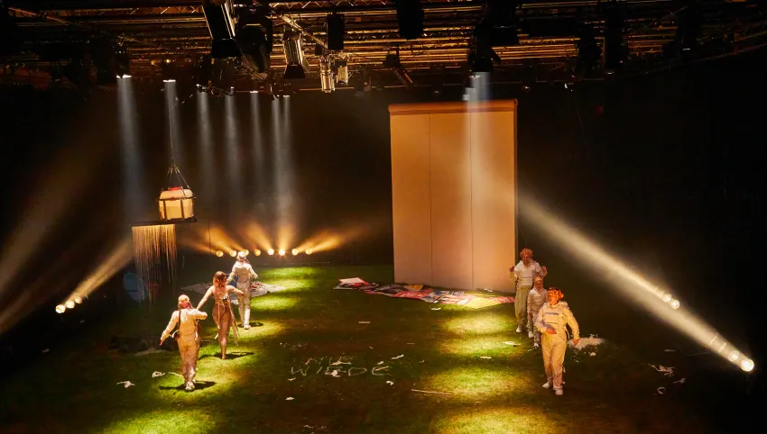 Bühnenbild mit grünem Boden, sechs Personen in hellen Kostümen, Scheinwerferlicht und einer großen weißen Wand im Hintergrund.