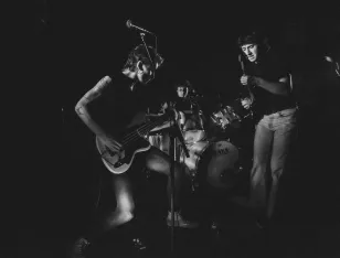 Drei Personen mit Schlagzeug und E-Gitarren.