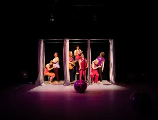 Ein Gymnastikball liegt vor sieben Personen, die sich zwischen Gardinen befinden. Eine von ihnen steht mit Gitarre vor einem Mikrofon.