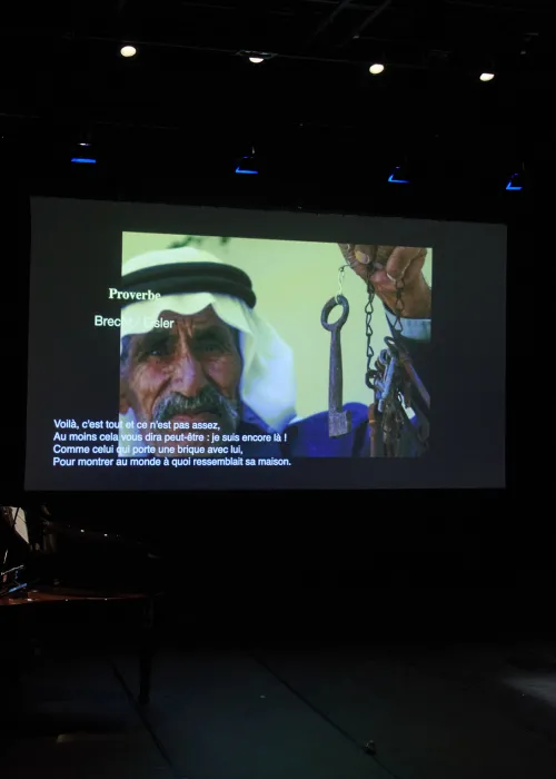 An der Wand ist eine Projektion von einem Mann, der einen alten Schlüsselbund in der Hand hat. Auch steht auf dem Bild: "Proverbe. Brecht / Eisler. Voilà, c'est tout et ce n'est pas assez, Au moins cela vous dira peut-être : je suis encore là ! Comme celui qui porte une brique avec lui, Pour montrer au monde à quoi ressemblait sa maison." Außerdem sitzen zwei Personen an einem Tisch, auf dem ein Laptop und mehrere Papierblätter liegen.