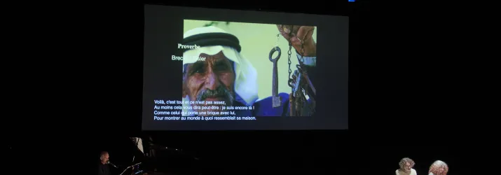 An der Wand ist eine Projektion von einem Mann, der einen alten Schlüsselbund in der Hand hat. Auch steht auf dem Bild: "Proverbe. Brecht / Eisler. Voilà, c'est tout et ce n'est pas assez, Au moins cela vous dira peut-être : je suis encore là ! Comme celui qui porte une brique avec lui, Pour montrer au monde à quoi ressemblait sa maison." Außerdem sitzen zwei Personen an einem Tisch, auf dem ein Laptop und mehrere Papierblätter liegen.