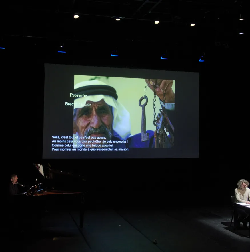 An der Wand ist eine Projektion von einem Mann, der einen alten Schlüsselbund in der Hand hat. Auch steht auf dem Bild: "Proverbe. Brecht / Eisler. Voilà, c'est tout et ce n'est pas assez, Au moins cela vous dira peut-être : je suis encore là ! Comme celui qui porte une brique avec lui, Pour montrer au monde à quoi ressemblait sa maison." Außerdem sitzen zwei Personen an einem Tisch, auf dem ein Laptop und mehrere Papierblätter liegen.