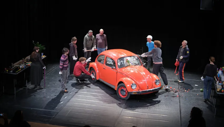 Ein roter VW Käfer, umgeben von mehreren Personen. Eine davon schlägt die Scheibe ein.