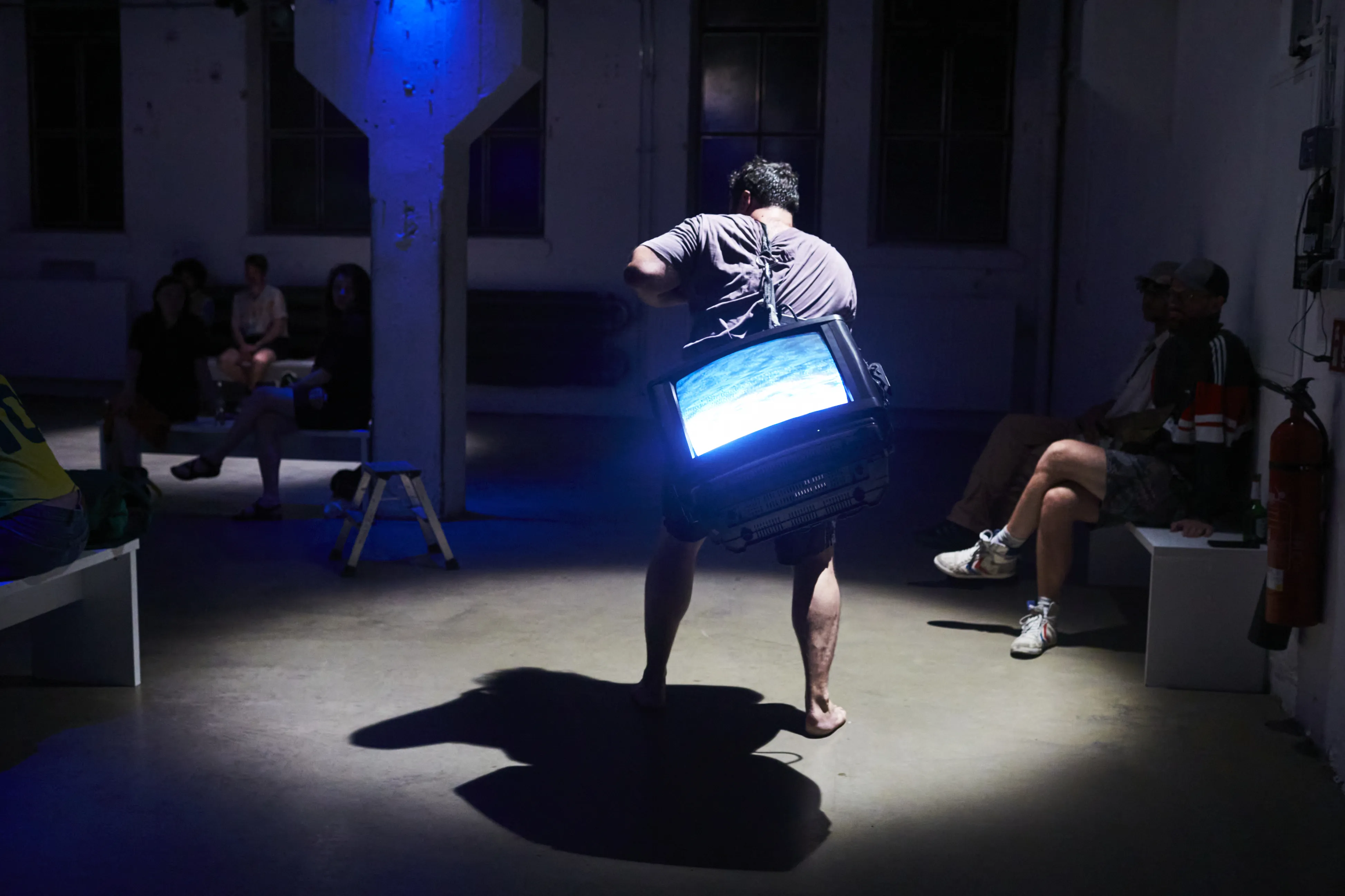 Eine Person, die einen Fernseher auf ihrem Rücken trägt und barfuß durch einen Raum läuft. An der Seite sitzen andere Menschen.