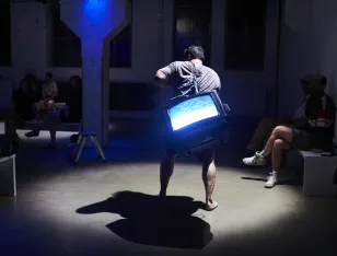 Eine Person, die einen Fernseher auf ihrem Rücken trägt und barfuß durch einen Raum läuft. An der Seite sitzen andere Menschen.