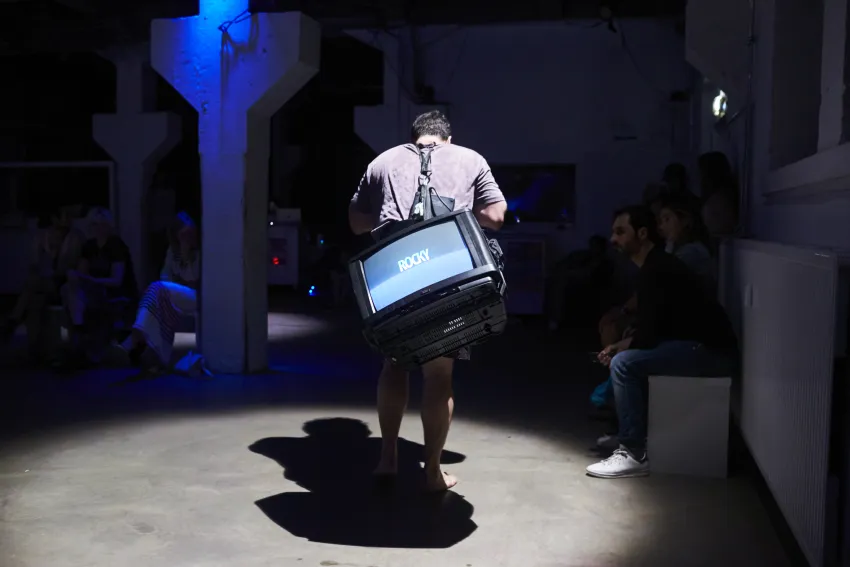 Eine Person, die einen Fernseher auf ihrem Rücken trägt, auf dem "Rocky" steht. An der Seite sitzen andere Menschen.