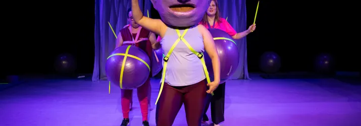 Eine Person mit einer großen Maske auf dem Kopf und einem gelben Stab in der Hand. Hinter ihr stehen zwei weitere Personen mit Gymnastikbällen um ihre Bäuche.