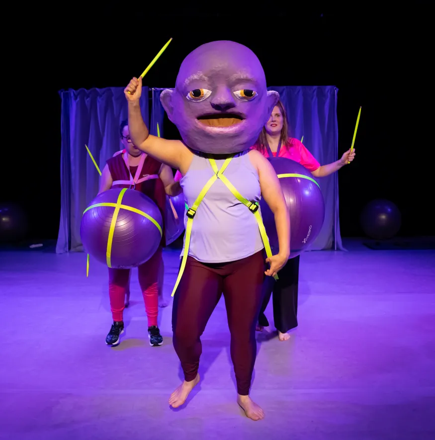 Eine Person mit einer großen Maske auf dem Kopf und einem gelben Stab in der Hand. Hinter ihr stehen zwei weitere Personen mit Gymnastikbällen um ihre Bäuche.