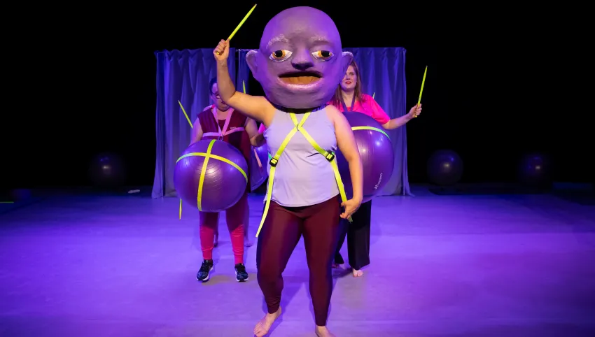 Eine Person mit einer großen Maske auf dem Kopf und einem gelben Stab in der Hand. Hinter ihr stehen zwei weitere Personen mit Gymnastikbällen um ihre Bäuche.