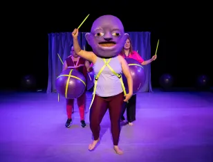 Eine Person mit einer großen Maske auf dem Kopf und einem gelben Stab in der Hand. Hinter ihr stehen zwei weitere Personen mit Gymnastikbällen um ihre Bäuche.