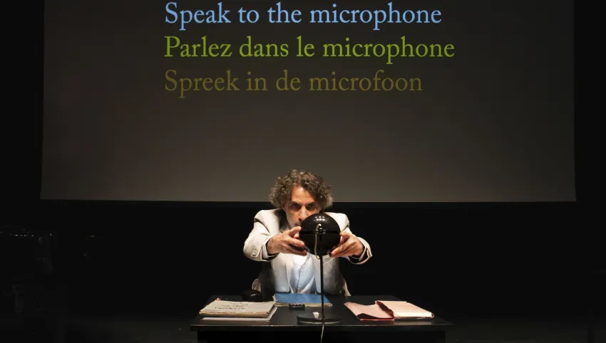 Eine Person an einem Tisch. Hinter ihr steht „Speak to the microphone. Parlez dans le microphone. Spreek in de microfoon.”