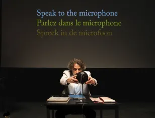 Eine Person an einem Tisch. Hinter ihr steht „Speak to the microphone. Parlez dans le microphone. Spreek in de microfoon.”