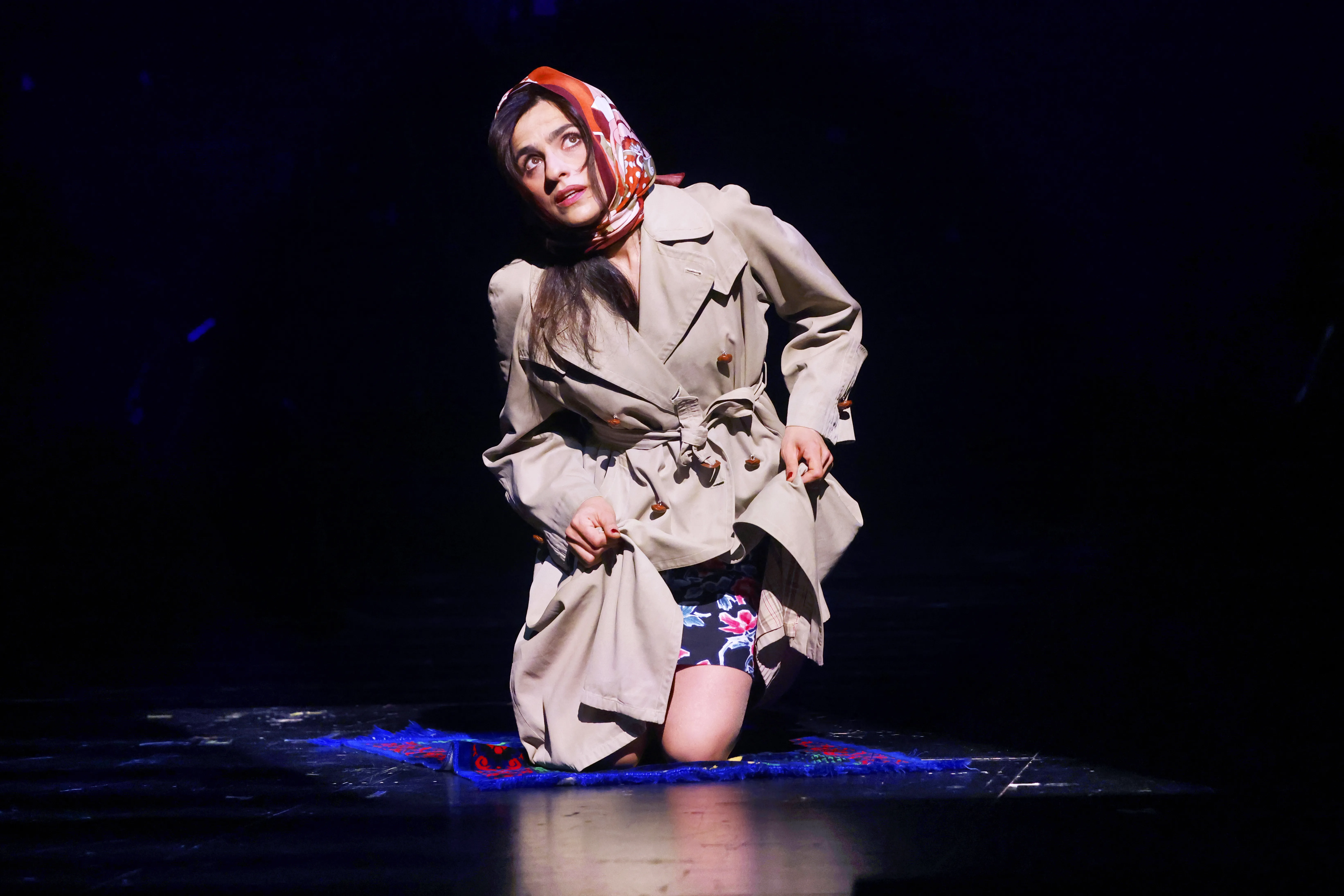 Eine Frau kniet und blickt nach oben. Sie trägt einen beigen Mantel und ein rotes Kopftuch.