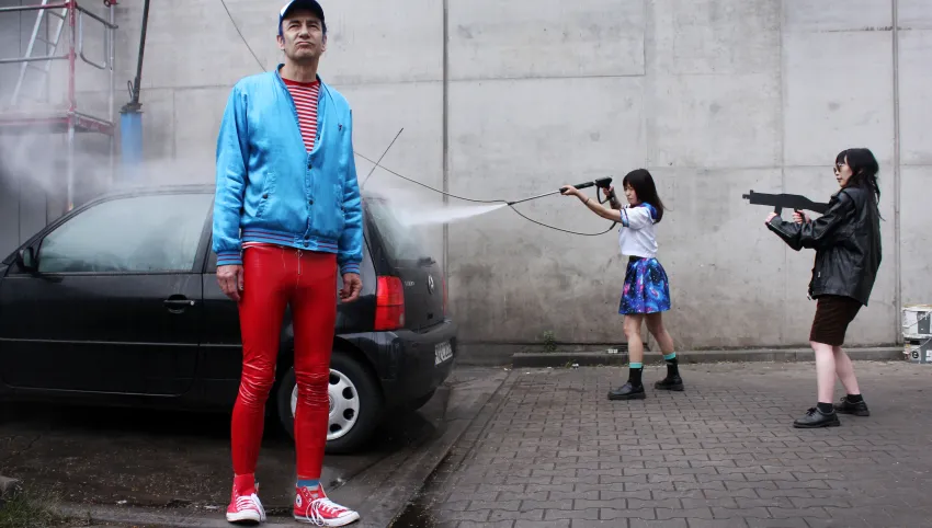 Eine Person steht vor einem schwarzen Auto. Zwei Personen spritzen Wasser auf das Auto.