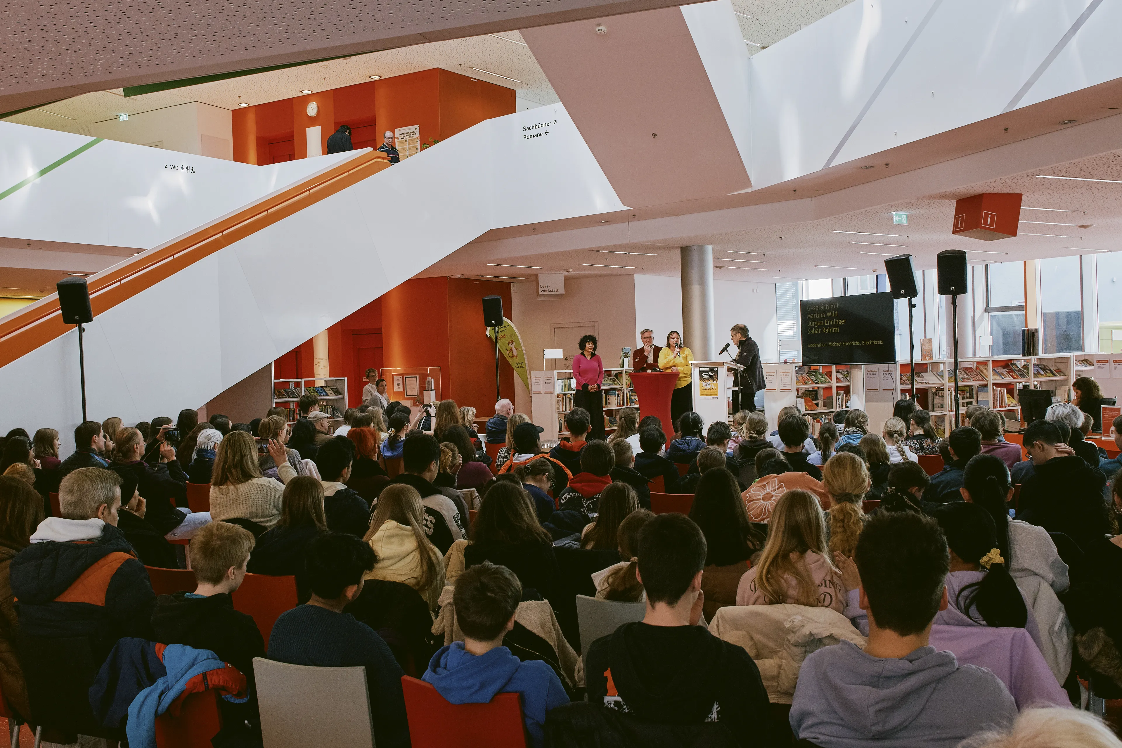 Im Foyer der Stadtbücherei Augsburg sind 150 Schüler versammelt. Der Brechtexperte Michael Friedrichs spricht auf dem Podium mit Sahar Rahami (Co-Festivalleitung), Jürgen Enninger (Kulturreferent) und Martina Wild (Bildungsreferentin) über die Bedeutung des Kreativwettbewerbs beim Brechtfestival