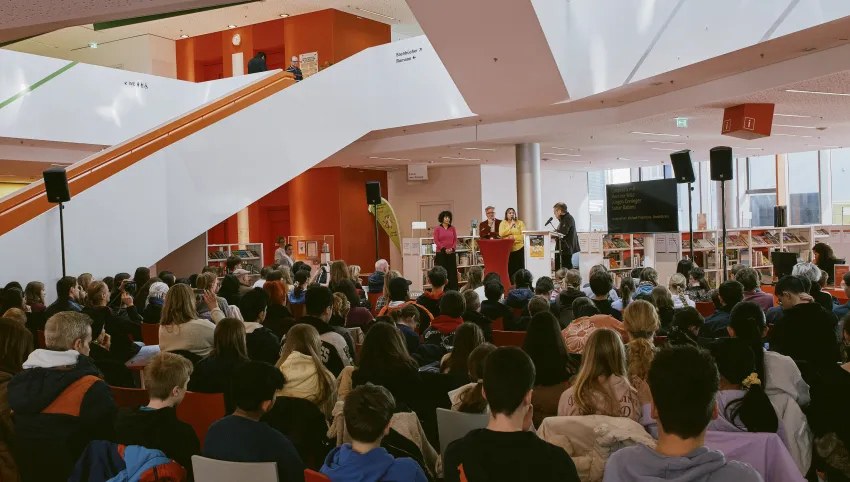 Im Foyer der Stadtbücherei Augsburg sind 150 Schüler versammelt. Der Brechtexperte Michael Friedrichs spricht auf dem Podium mit Sahar Rahami (Co-Festivalleitung), Jürgen Enninger (Kulturreferent) und Martina Wild (Bildungsreferentin) über die Bedeutung des Kreativwettbewerbs beim Brechtfestival
