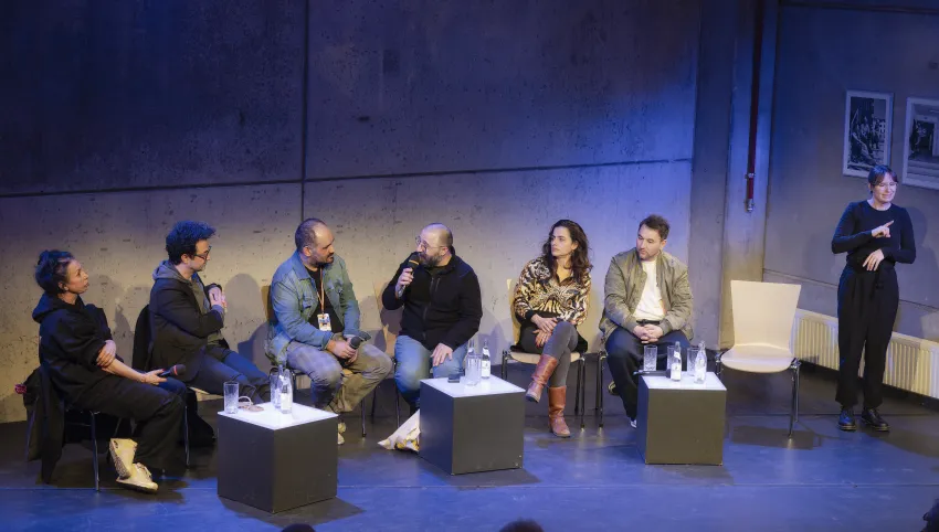 Der Autor Dinçer Güçyeter sitzt umrahmt von Künstlerinnen des Maxim Gorki Theaters. Düzgün Polat stellt ihm eine Frage zu seinem Roman "Unser Deutschlandmärchen". Eine DGS Dolmetscherin übersetzt das Gespräch.
