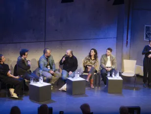Der Autor Dinçer Güçyeter sitzt umrahmt von Künstlerinnen des Maxim Gorki Theaters. Düzgün Polat stellt ihm eine Frage zu seinem Roman "Unser Deutschlandmärchen". Eine DGS Dolmetscherin übersetzt das Gespräch.
