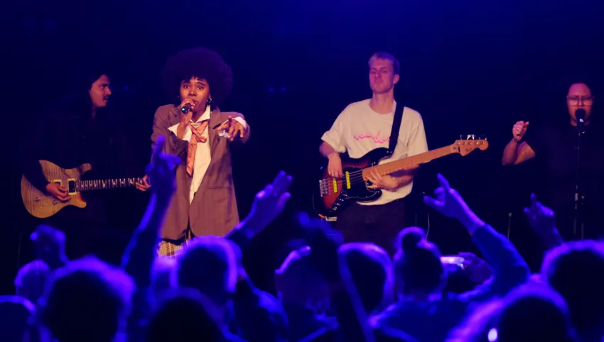 Sorvina singt uns streckt den linken Arm nach dem Publium aus. Das Publikum winkt ihr zu. Neben Sorvina spielt ein Mann im weißen T-Shirt Gitarre
