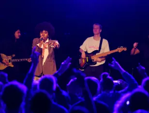 Sorvina singt uns streckt den linken Arm nach dem Publium aus. Das Publikum winkt ihr zu. Neben Sorvina spielt ein Mann im weißen T-Shirt Gitarre