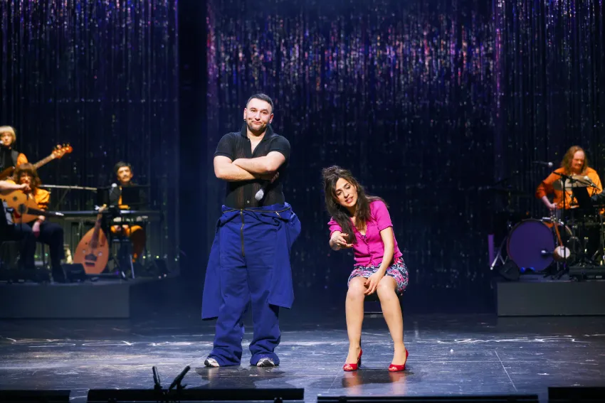 Ein Mann mit verschränkten Armen steht neben einer sitzenden Frau.