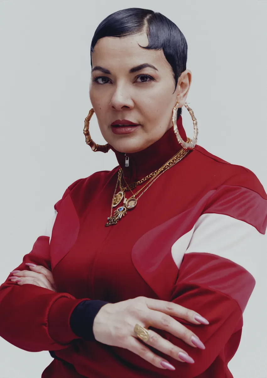 Eine Person in roter Kleidung und goldenem Schmuck, die Arme verschränkt.