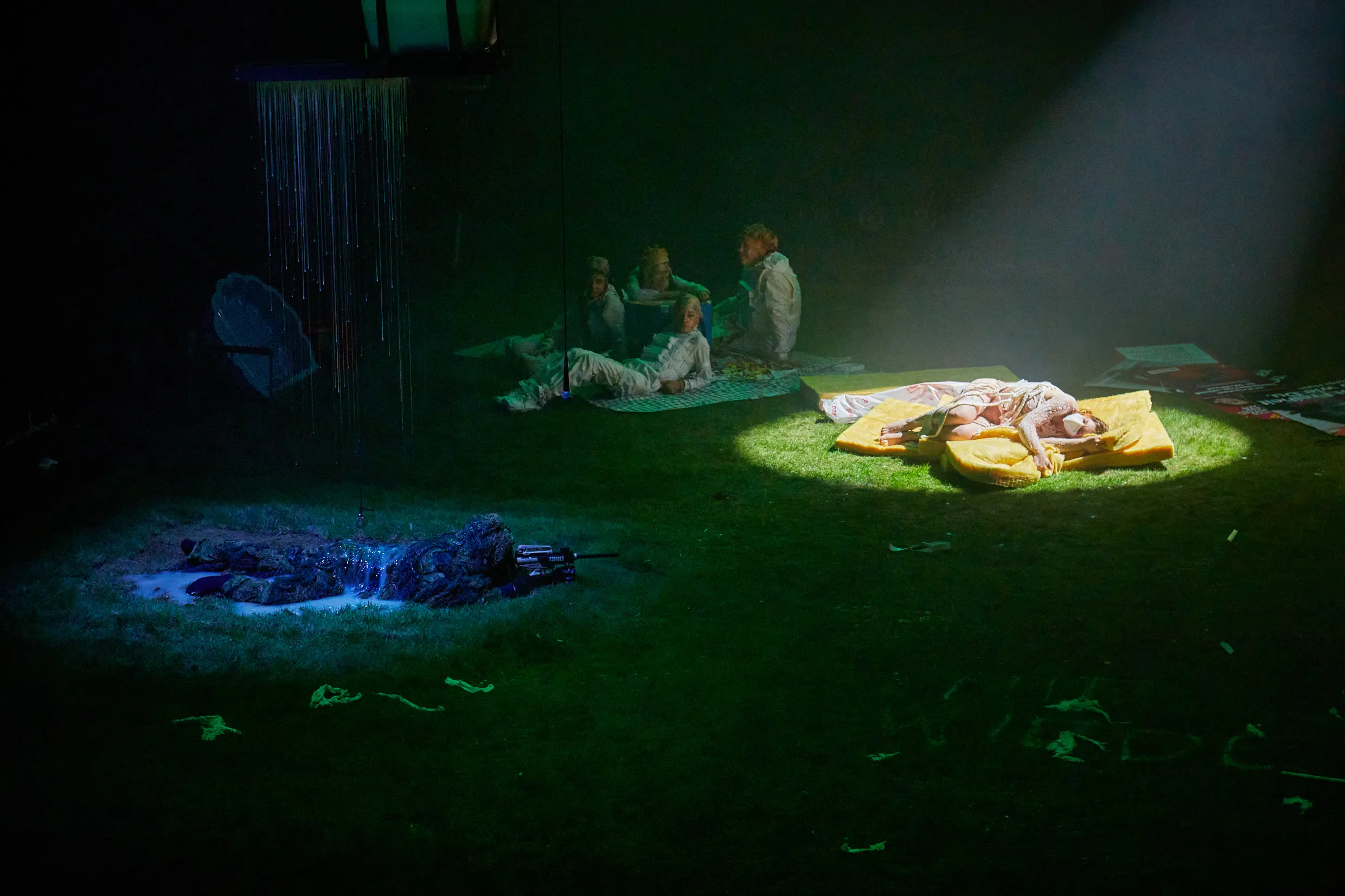 Mehrere Personen liegen auf grünem Boden, wovon eine mit Scheinwerferlicht beleuchtet wird.